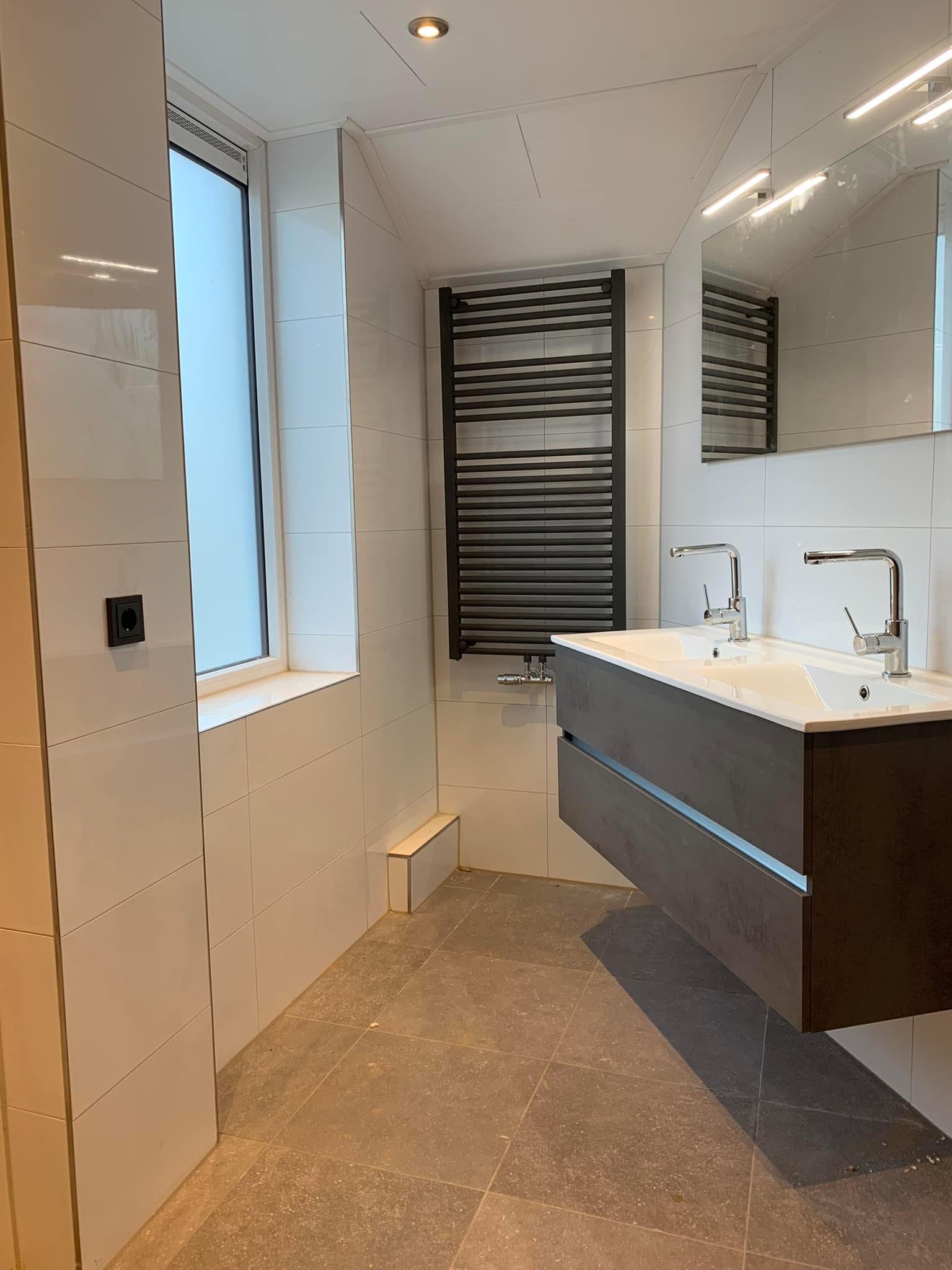Lichte badkamer met zwarte radiator en dubbele wastafel, betegeld door Van der Wal Tegelwerk